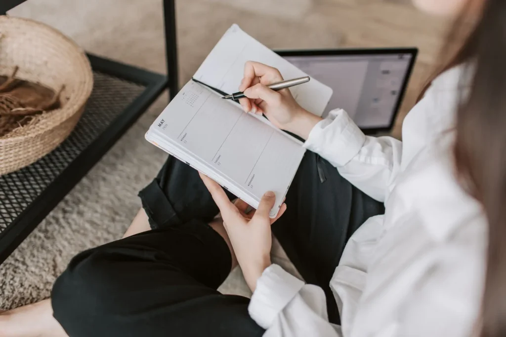 A young woman sitting on the floor at home, writing in a planner for a wedding with a pen
