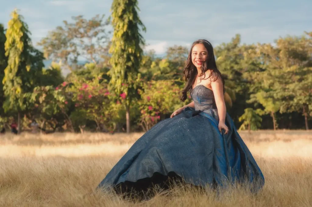 A teenager in a flowing blue dress stands gracefully in a vibrant green field under a clear blue sky.